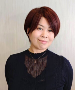 Portrait of a woman with short burgundy hair, wearing a black knit top and a delicate necklace, smiling at the camera.