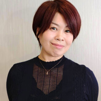 Portrait of a woman with short burgundy hair, wearing a black knit top and a delicate necklace, smiling at the camera.